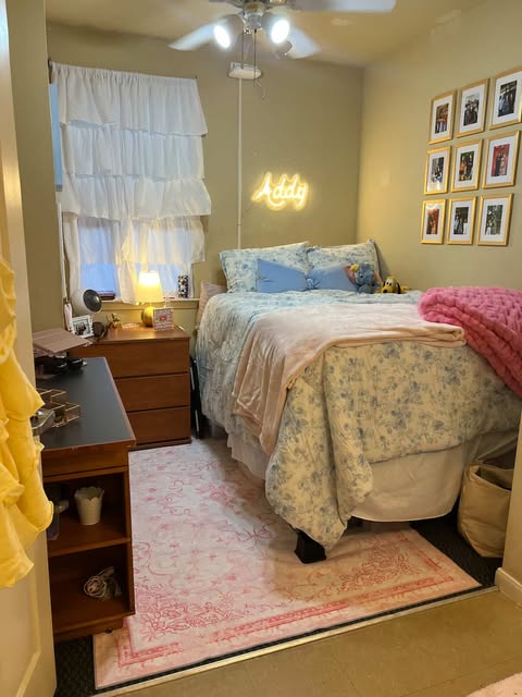 A cozy bedroom with a neatly made bed featuring floral bedding, a wall decorated with framed photographs, and a neon sign that reads